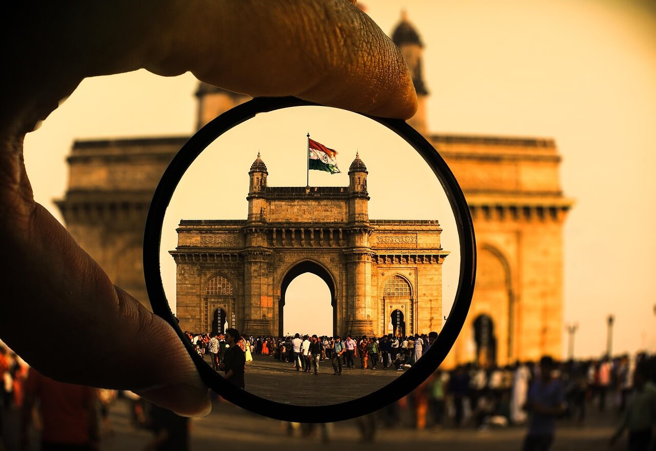 A hand holding a circular camera filter framing the Gateway of India with the Indian flag on top and a crowd of visitors below at sunset.