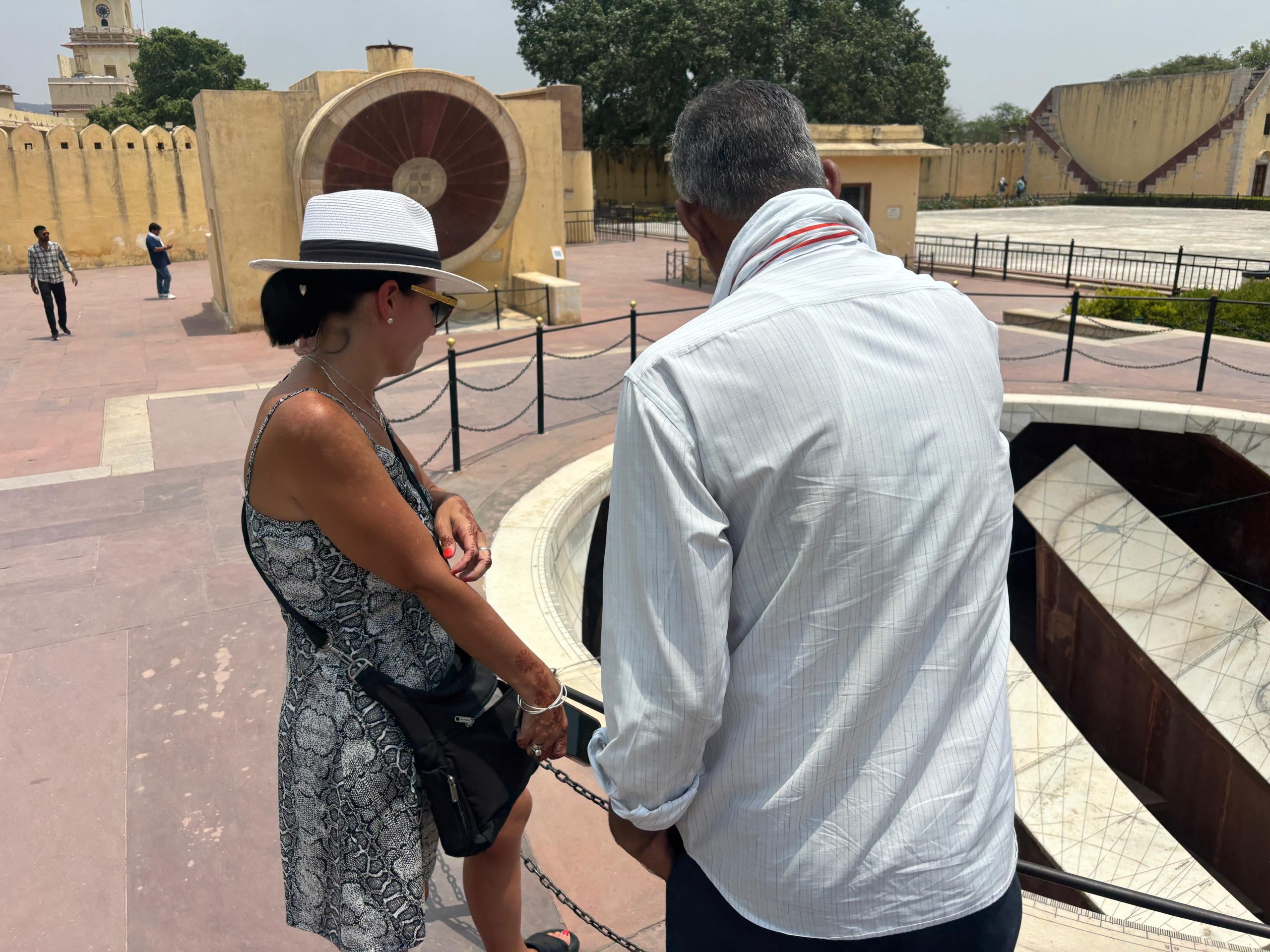 Two people stand near an ancient astronomical instrument at an outdoor observatory with sandstone structures and geometric markings. The woman wears a white hat and patterned dress, while the man wears a light shirt. The scene is bright and sunny, with other visitors and historic architecture visible in the background.