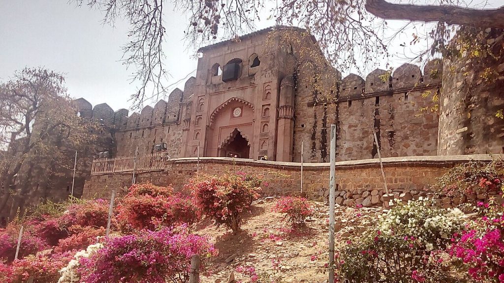 The historic Jhansi Fort entrance, a symbol of Rani Lakshmibai’s legacy and 18th-century military architecture. Jhansi Fort stone walls and arched entrance with flowers in foreground