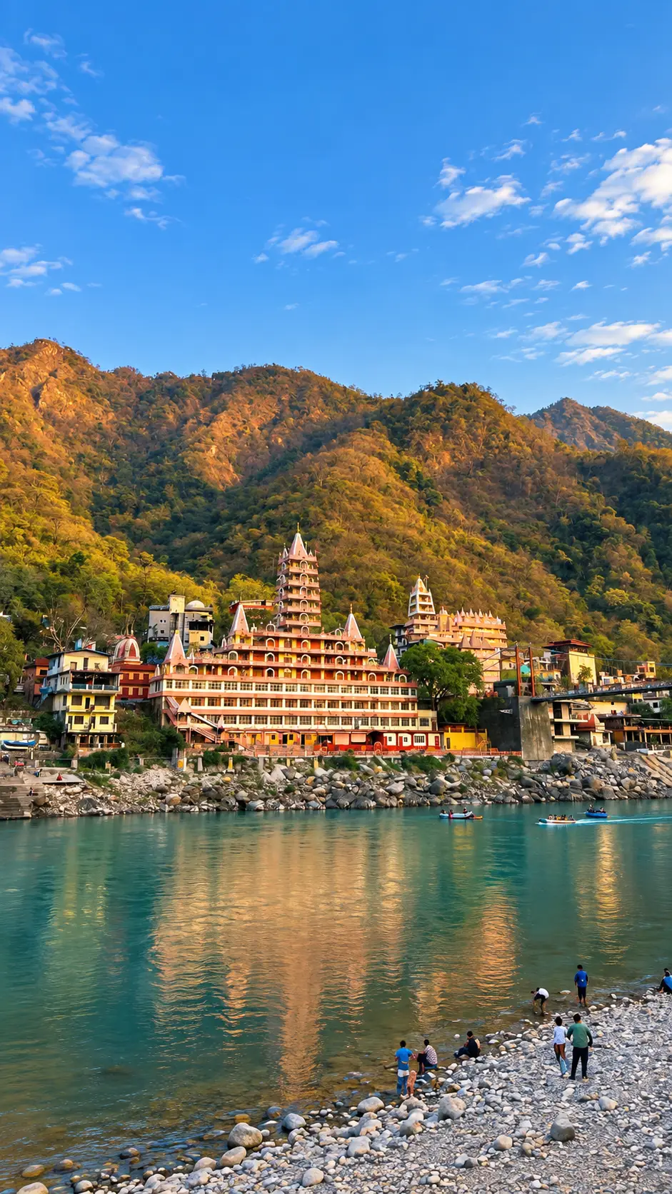 Riverside temple buildings and boats along the Ganges River in Rishikesh, India, with forested hills in the background.