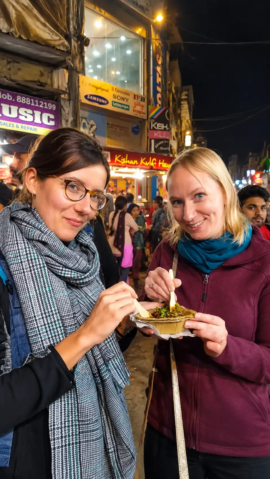 Two female tourists enjoying Indian chaat at a busy night market in Agra.